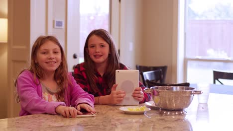 Two-friends-sitting-at-a-kitchen-island-using-their-tablets-and-smiling-at-the-camera