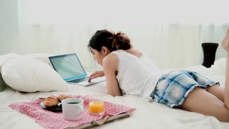 Young-woman-using-laptop-during-breakfast-lying-on-white-bed-at-home.-Brunette-girl-typing-on-pc-computer
