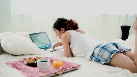 Joven-usando-laptop-durante-el-desayuno-de-mentira-en-cama-blanca-en-casa.-Chica-morena-escribiendo-en-la-computadora-de-la-pc