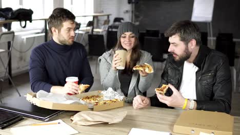 Eine-Gruppe-von-jungen-Designern-geführt-durch-den-Leiter-arbeiten-an-dem-Projekt-von-Design-Büro-und-essen-Pizza-und-trinken-Tee-oder-Kaffee-im-Pappbecher-geliefert.-Schuss-in-4k