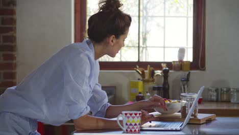 Mujer-joven-en-pijama-usando-laptop-en-la-cocina-comiendo-fruta,-disparos-a-R3D