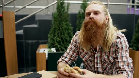 Estudiante-barbudo-hombre-comer-hamburguesa-en-la-calle-de-la-cafetería-al-aire-libre