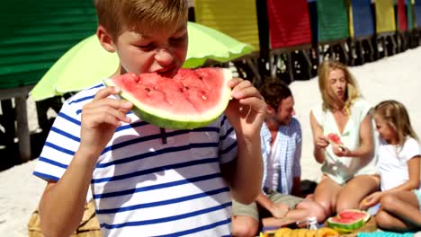 Junge-Essen-Wassermelone-während-Familie-sitzt-im-Hintergrund-am-Strand