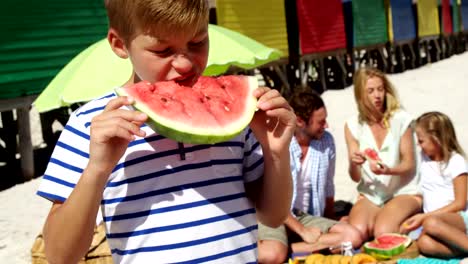 Boy-eating-watermelon-while-family-sitting-in-background-at-beach