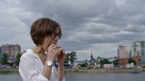 Businesswoman-Walking-along-Riverside-and-Chewing-Burger