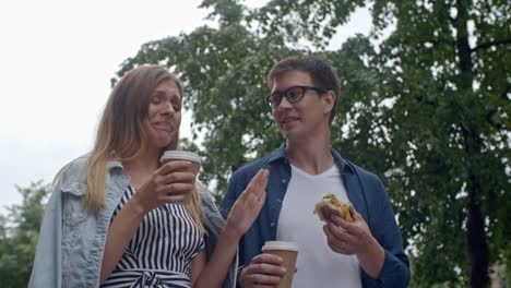 Joven-pareja-comer-hamburguesas-y-beber-café-durante-caminata