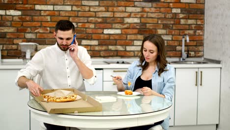 Mann-und-schwangere-Frau-Essen-Pizza-zu-Hause-in-ihrer-Küche.-Mann-am-Telefon-zu-sprechen,-während-des-Abendessens