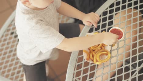 Downward-shot-of-a-little-kid-dipping-his-curly-fry-in-ketchup-and-eats-it-at-an-outdoor-food-court