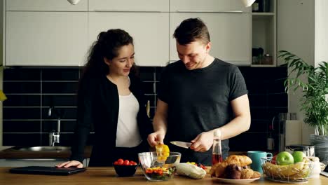 young-beautiful-caucasian-pretty-curly-businesswoman-came-home-from-work,-her-handsome-husband-is-cooking,-cutting-yellow-pepper,-they-are-kissing-and-laughing