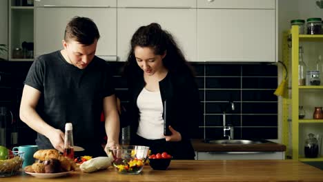 joven-empresaria-bella-llegó-a-casa-del-trabajo-ah-Inicio,-le-apuesto-marido-está-cocinando-en-la-cocina,-ella-pregunta-sobre-cena,-besándose-y-riendo