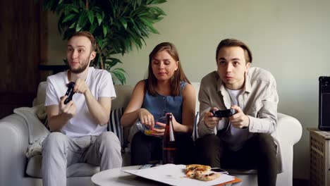 Two-male-friends-are-playing-video-games-pressing-buttons-on-joystick-while-pretty-blond-girl-is-watching-game-and-eating-snacks.-Friendship-and-technology-concept.