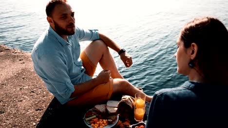 Young-happy-couple-sitting-on-the-shore-of-a-sea-and-eating.-Man-and-woman-having-picnic-near-water-in-the-morning