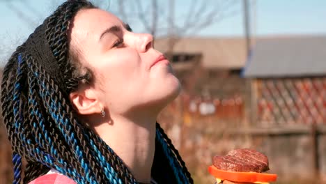 Young-woman-with-blue-braid-hairs-eats-sandwich-with-bread,-cutlet,-pepper-and-cheese-outdoor.