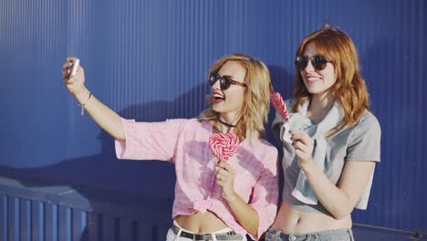 Dos-mujeres-jóvenes-atractivas-en-gafas-de-sol,-hablar,-comer-Chupa-Chups-y-tomar-un-selfie-en-calle