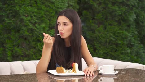 Un-dolly-shot-de-una-morena-guapa-joven-comiendo-un-pastel-sentado-afuera.-Trata-de-una-sola-pieza-con-deleite,-mira-a-la-izquierda-y-sigue-comiendo