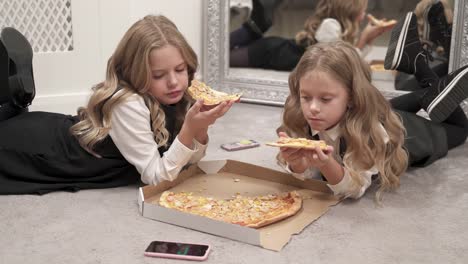 Zwei-ziemlich-blonde-Mädchen-liegen-auf-dem-Boden-essen-Pizza.-Es-ist-eine-geöffnete-Box-mit-Pizza-vor-ihnen.-Sie-blicken-auf-den-Handy-Bildschirm.