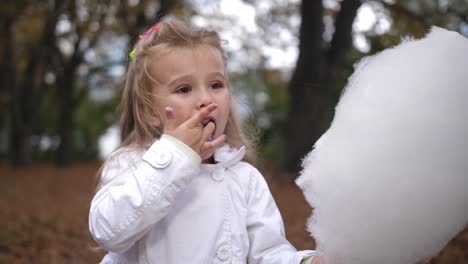 Niña-rubia-come-dulce-algodón-de-azúcar-en-el-parque-de-la-ciudad.-Hermosa-niña-comiendo-algodón-de-azúcar.-Niño-comiendo-algodón-de-azúcar-verde-bosque-en-el-fondo.-Lenta-de-4-k.