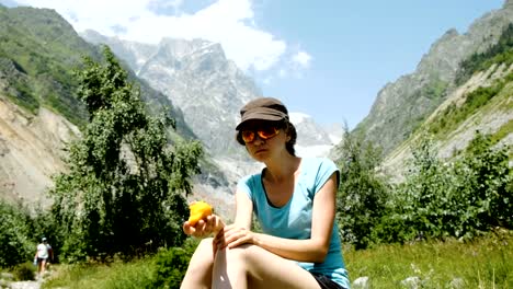 Woman-tourist-sits-on-a-stone-and-eats-a-peach-in-a-hike-on-the-background-of-a-beautiful-mountain-landscape