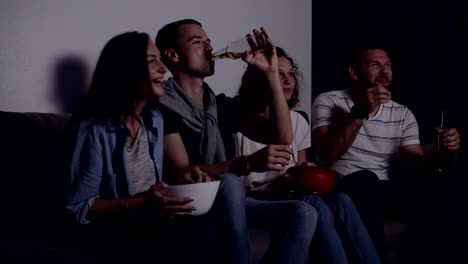 Friends-of-four-sit-on-couch,-watch-funny-film-on-TV.-Caucasian-young-group-sit-on-the-couch,-drinks-and-snacks.-Movie-night