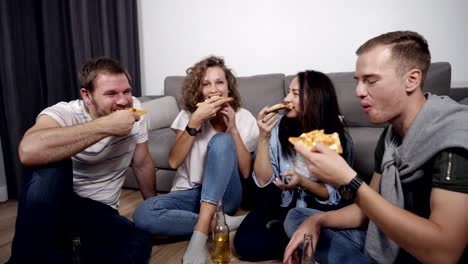 Group-of-four-friends-having-fun,-ordered-large-pizza-and-eating,-laughing-and-talking,-sitting-on-floor-in-grey-and-white-coloured-loft-room.-Drinking-and-spending-time-together