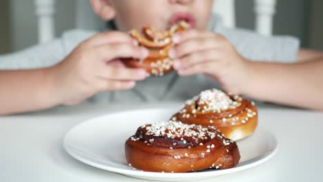 Fröhlicher-Junge-sitzt-hinter-dem-Tisch-und-Essen-eine-Zimtschnecke,-Nahaufnahme