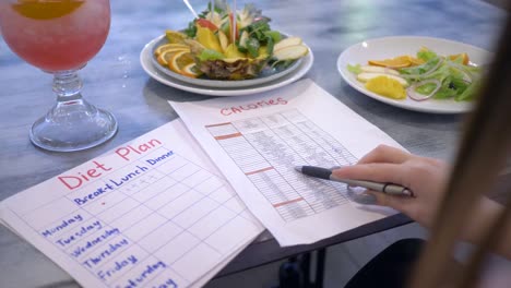 mujer-de-nutricionista-buscando-calorías-en-lista-y-sostenga-en-la-mano-sentado-en-la-mesa-con-frutas-en-placa-y-punch-de-plan-de-dieta