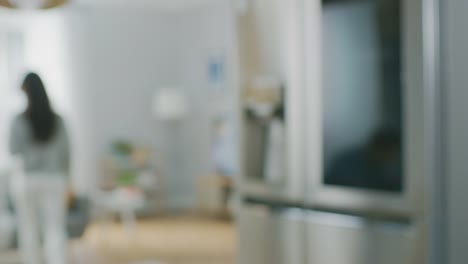 Beautiful-Brunette-Girl-in-White-Jeans-and-Grey-Sweater-Comes-to-the-High-Tech-Fridge-with-a-Glass-Door-and-Takes-a-Green-Apple.-She-goes-to-the-Living-Room.-Flat-has-Modern-Interior.