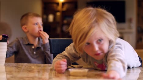 Una-niña-que-alcanza-para-la-comida-en-el-mostrador-de-la-cocina,-con-su-hermano-en-el-fondo