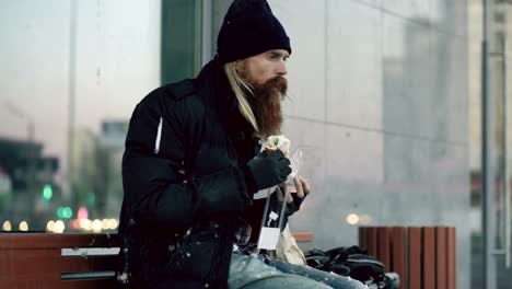 Homeless-young-man-eating-sandwich-on-bench-at-city-street-in-evening