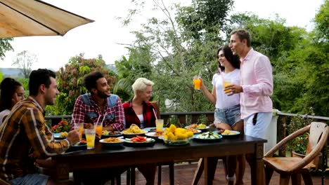 Par-decir-brindis-de-pie-gente-comiendo-en-la-terraza-jóvenes-amigos-sentados-en-la-mesa-al-aire-libre-comunicación