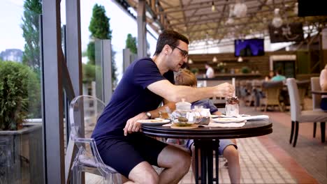 Gut-aussehender-Mann-in-lässigen-blaues-Hemd-und-Hosen-sitzen-im-Restaurant-mit-Sohn-und-Menschen-im-Hintergrund.-Junger-Vater-Essen-Dessert-des-Kindes-mit-langen-Teelöffel.-Kleiner-Junge-im-Gespräch-mit-männlicher-Elternteil