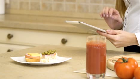 Girl-eats-sandwich-and-uses-tablet-standing-at-the-kitchen