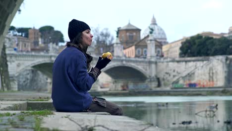 junge-Obdachlose-einsame-Frau-sitzen-am-Fluss-isst-einen-Apfel