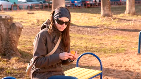 Junge-Brünette-Frau-mit-Sonnenbrille-schält-und-isst-Mandarine-sitzen-auf-der-Bank-im-Park.