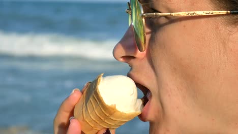 Young-brunette-woman-licking-eating-icecream-on-the-beach,-blue-sea-background-and-sunglasses.