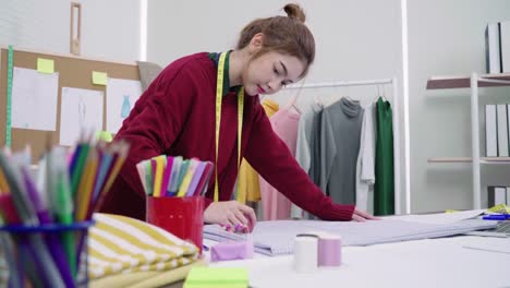 Junge-asiatische-Frau-Mode-Designer-Zeichnung-mit-Bleistift-und-Papier-während-der-Arbeit-im-Workshop-Studio-betrachten.