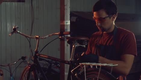 theme-small-business-bike-repair.-A-young-Caucasian-brunette-man-wearing-safety-goggles,-gloves-and-an-apron-uses-a-hand-tool-to-repair-and-adjust-the-bike-in-the-workshop-garage