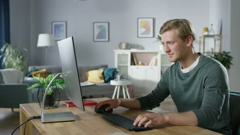 Portrait-of-the-Focused-Young-Man-Working-on-a-Personal-Computer-while-Sitting-at-His-Desk.-In-the-Background-Cozy-Living-Room.