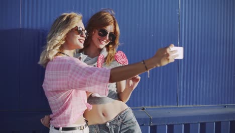 Two-young-attractive-women-in-sun-glasses-talking,-eat-lollipop-and-take-a-selfie-on-street