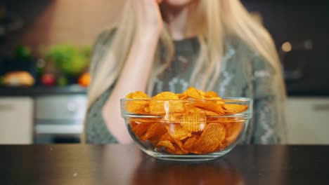 Froh,-dass-Frau-essen-Kartoffelchips.-Schöne-junge-Frau-genießen-Kartoffelchips-und-Blick-in-die-Kamera-während-der-Sitzung-in-stilvolle-Küche