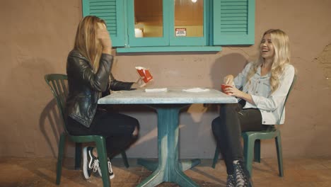 Two-girls-sitting-at-table-outside-playing-with-food-throwing-it-at-each-other-laughing