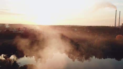 Rauch-vom-Feuer-breiten-sich-über-den-Fluss-bei-Sonnenuntergang-auf-dem-Hintergrund-eines-Kraftwerks-mit-zwei-Rohren.