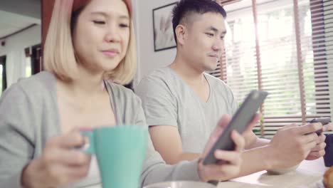 Attractive-young-Asian-couple-distracted-at-table-with-newspaper-and-cell-phone-while-eating-breakfast.-Excited-young-Asian-couple-surprised-by-unbelievably-good-news,-happy-family-amazed-by-internet.