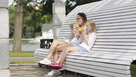 Una-mujer-joven-con-una-chica-rubia-y-descansar-en-el-parque-y-comer-maíz-caliente.