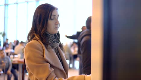 Young-brunette-woman-in-coat-orders-food-through-terminal
