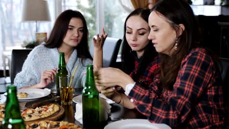 Alegre-compañía-de-las-niñas-en-el-restaurante-viendo-fotos-en-el-smartphone.-Chicas-comer-pizza,-beber-cerveza,-charlar-y-divertirse.