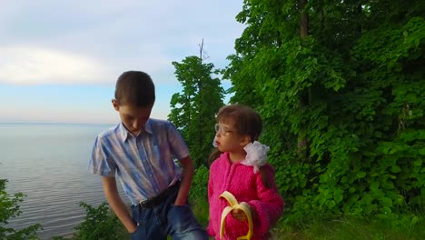 Children-have-a-rest-in-the-forest-on-the-banks-of-a-large-lake.-Girl-child-eats-a-banana.