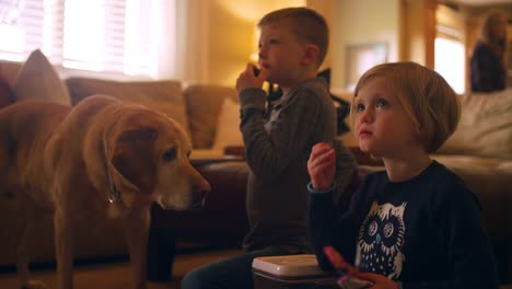 Siblings-watching-television-together-while-eating-snacks,-with-the-family-dog