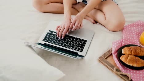 Young-woman-using-laptop-during-breakfast-sitting-on-white-bed-at-home.-Close-up-view-of-girl-typing-on-pc-computer