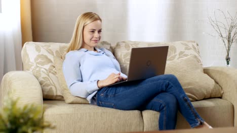 Hermosa-mujer-rubia-sentada-en-un-sofá-con-el-portátil-en-su-regazo,-ella-tipos-activamente-en-él.-Su-cuarto-está-lleno-de-luz.